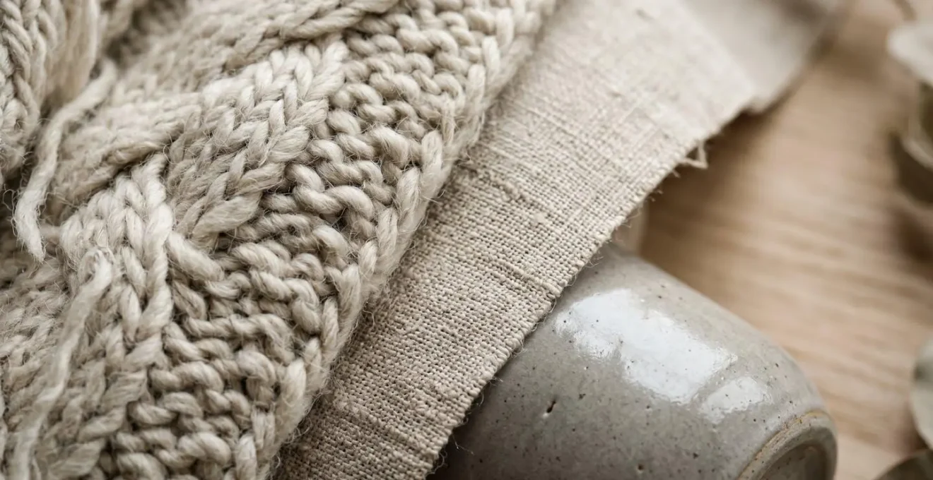 Close-up composition of layered neutral textures including raw linen fabric, chunky wool knit, smooth ceramic, and natural wood grain